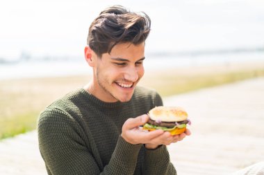 Açık havada elinde hamburgerle dolaşan beyaz bir genç.