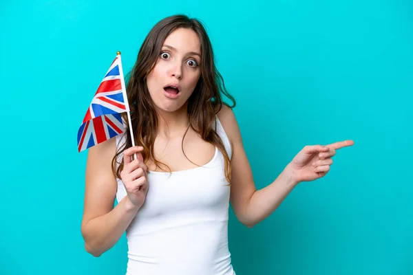 年轻的高加索女人拿着英国国旗 蓝色背景孤立 惊讶地指向一边 — 图库照片