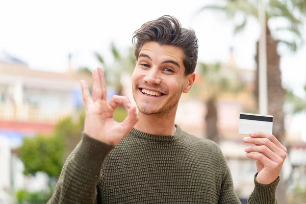 Młody Biały Mężczyzna Trzyma Kartę Kredytową Zewnątrz Pokazując Znak Palcami — Zdjęcie stockowe