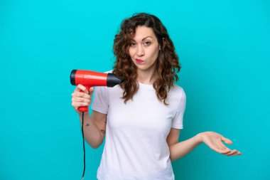 Mavi arka planda saç kurutma makinesi tutan beyaz bir kadın, omuzlarını kaldırırken şüpheyle hareket ediyor.