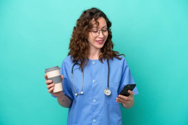 Young nurse caucasian woman isolated on blue background holding coffee to take away and a mobile