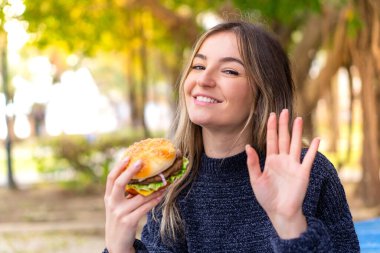 Genç ve güzel Romen kadın elinde hamburgerle elini sallayarak neşeli bir ifadeyle selam veriyor.