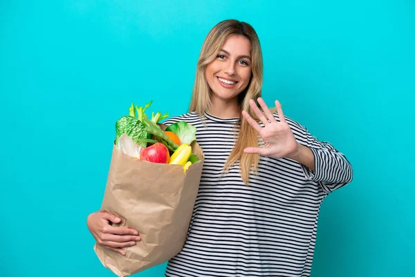 Young Uruguayan woman holding a grocery shopping bag isolated on blue background counting five with fingers