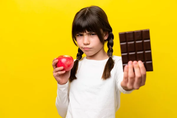 Little caucasian girl isolated on yellow background taking a chocolate tablet in one hand and an apple in the other