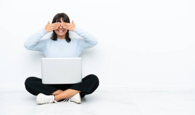 Genç melez kadın dizüstü bilgisayarla yerde oturuyor. Beyaz arka planda izole edilmiş. Gözlerini elleriyle kapatıyor.
