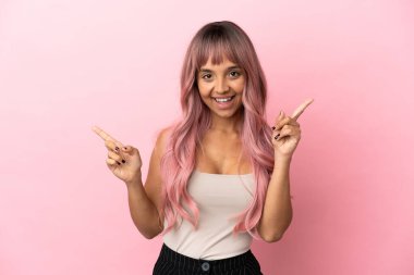Pembe saçlı genç melez kadın pembe arka planda izole edilmiş parmak uçlarını işaret ediyor ve mutlu.