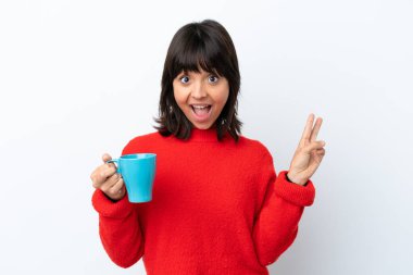 Beyaz arka planda izole edilmiş bir fincan kahve tutan beyaz kadın gülümsüyor ve zafer işareti gösteriyor.