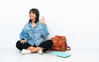 Genç bir kız öğrenci, beyaz arka planda tek başına oturuyor. Parmaklarıyla işaret veriyor.