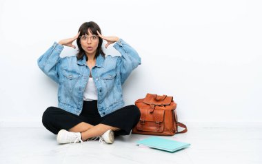 Genç bir kız öğrenci, beyaz bir zeminde oturuyor, şaşırmış bir şekilde.