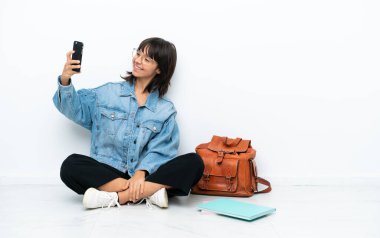 Genç bir kız öğrenci, beyaz arka planda izole bir şekilde yerde oturuyor ve selfie çekiyor.