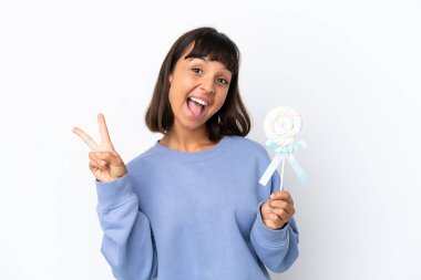 Beyaz arka planda izole edilmiş lolipop tutan genç bir ırk kadını gülümsüyor ve zafer işareti gösteriyor.