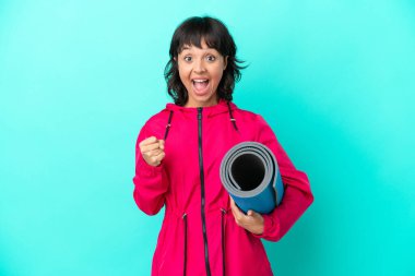 Mavi arka planda bir minder tutarak yoga derslerine giden genç kız galibiyetini kutluyor.