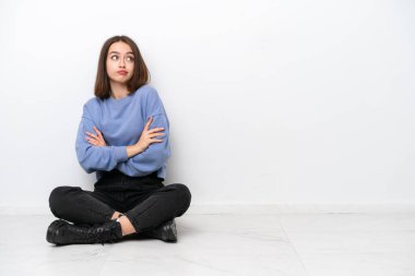 Genç Ukraynalı kadın, beyaz arka planda izole bir şekilde yerde oturuyor. Omuzlarını kaldırırken şüpheyle hareket ediyor.