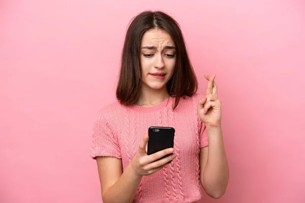Young Ukrainian woman isolated on pink background using mobile phone with fingers crossing