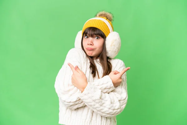 stock image Little caucasian girl wearing winter muffs over isolated background pointing to the laterals having doubts