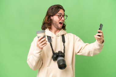 İzole edilmiş arka planda genç bir fotoğrafçı elinde kahve ve bir cep telefonu tutuyor.