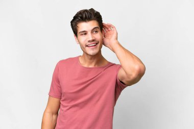 İzole edilmiş beyaz arka planda genç, yakışıklı beyaz bir adam elini kulağına koyarak bir şeyler dinliyor.