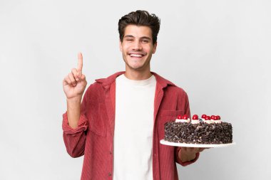 Kafkasyalı genç bir adam, izole edilmiş arka planda doğum günü pastası tutuyor ve en iyinin işareti olarak parmağını kaldırıyor.