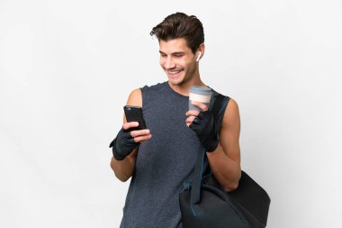 Young sport caucasian man with sport bag over isolated white background holding coffee to take away and a mobile