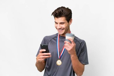 İzole edilmiş beyaz arka planda madalyaları olan genç beyaz bir adam elinde kahve ve bir cep telefonu tutuyor.