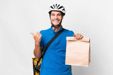 Young caucasian man taking a bag of takeaway food over isolated background pointing to the side to present a product
