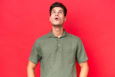Genç beyaz yakışıklı adam kırmızı arka planda izole edilmiş yukarı bakıyor ve şaşırmış bir ifadeyle