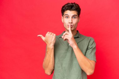 Kızıl arka planda izole edilmiş genç, beyaz, yakışıklı bir adam. Yan tarafı işaret ediyor ve sessiz hareketler yapıyor.