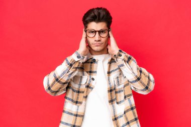 Kızıl arka planda izole edilmiş genç beyaz yakışıklı adam hayal kırıklığına uğramış ve kulaklarını kapatmış.