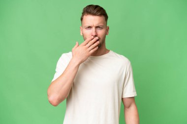 Sarışın, beyaz bir adam izole edilmiş arka planda esniyor ve ağzını eliyle kapatıyor.