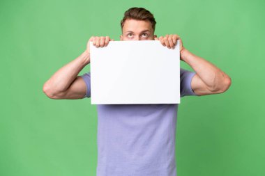 Sarışın, beyaz bir adam, izole edilmiş bir geçmişi var. Elinde boş bir levha var ve arkasına saklanıyor.