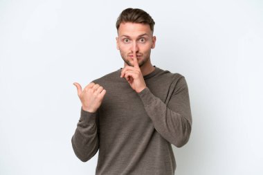 Beyaz arka planda izole edilmiş genç, sarışın, beyaz bir adam. Yan tarafı işaret ediyor ve sessizlik hareketi yapıyor.