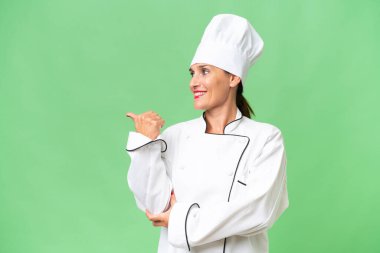 Orta yaşlı bir aşçı kadın, izole edilmiş arka planda bir ürün sunmak için kenarı işaret ediyor.