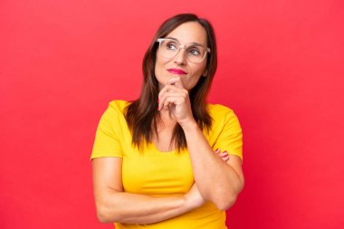 Orta yaşlı beyaz kadın, kırmızı arka planda izole edilmiş, yukarı bakarken bir fikir düşünüyor.