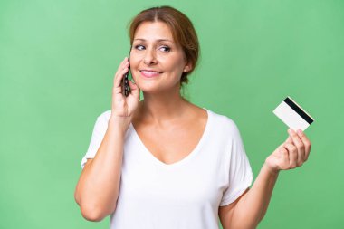 Orta yaşlı beyaz bir kadın izole edilmiş bir geçmişi var cep telefonuyla sohbet ediyor ve elinde bir kredi kartı tutuyor.