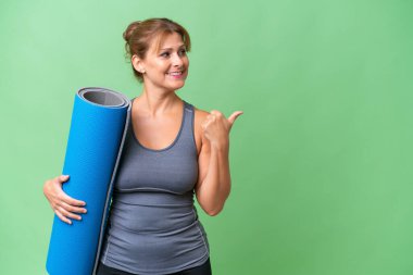 Orta yaşlı sporcu bir kadın yoga derslerine giderken elinde bir paspas tutarak bir ürünü sunmak için kenarı işaret ediyor.