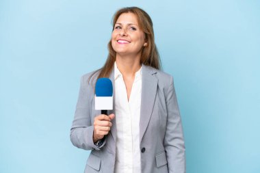 Orta yaşlı televizyon sunucusu kadın izole edilmiş mavi arka planda kolları kalçasında ve gülümsüyor.
