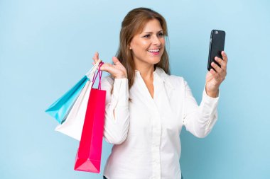 Orta yaşlı beyaz bir kadın mavi arka planda izole edilmiş. Elinde alışveriş poşetleri var ve bir arkadaşına cep telefonuyla bir mesaj yazıyor.