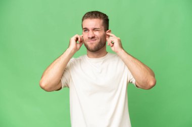 Yeşil krom arka planda izole edilmiş yakışıklı beyaz adam hayal kırıklığına uğramış ve kulaklarını kapatmış.