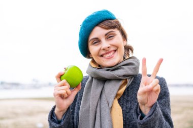 Açık havada elmalı esmer kadın gülümsüyor ve zafer işareti gösteriyor.