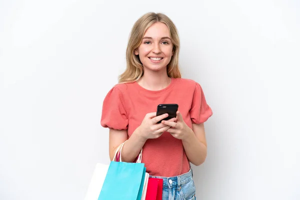 白い背景に隔離された若い英語の女性ショッピングバッグを保持し 友人に彼女の携帯電話でメッセージを書く — ストック写真