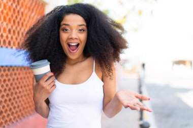 Genç Afrikalı Amerikalı bir kadın açık havada kahve tutuyor ve yüzünde şok ifadesi var.