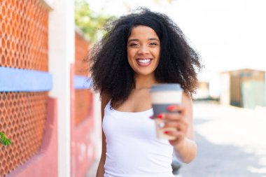 Genç Afro-Amerikalı kadın açık havada elinde kahveyle neşeli bir ifade tutuyor.