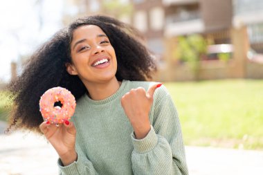 Genç Afrikalı Amerikalı bir kadın açık havada donut tutuyor ve bir ürün sunmak için kenarı işaret ediyor.