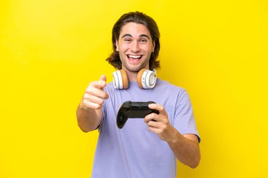 Sarı arka planda izole edilmiş bir bilgisayar oyunu kontrolörüyle oynayan yakışıklı beyaz adam şaşırmış ve ön tarafı işaret ediyor.