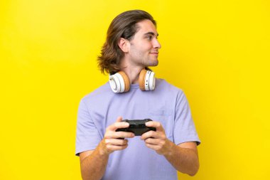 Sarı arka planda izole edilmiş bir bilgisayar oyunu kontrolörüyle oynayan yakışıklı beyaz adam yan tarafa bakıyor.