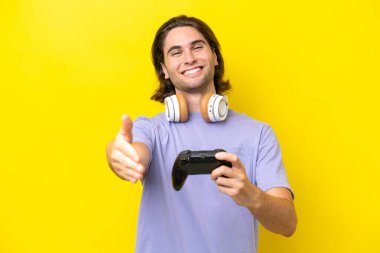 Sarı arka planda izole edilmiş bir bilgisayar oyunu kontrolörüyle oynayan yakışıklı beyaz adam iyi bir anlaşma için el sıkışıyor.