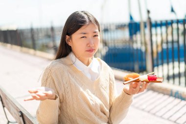 Genç Çinli bir kadın açık havada sashimi tutuyor. Omuzlarını kaldırırken şüpheyle hareket ediyor.