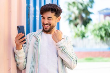 Yakışıklı bir Arap açık havada cep telefonu kullanıyor ve zaferi kutluyor.