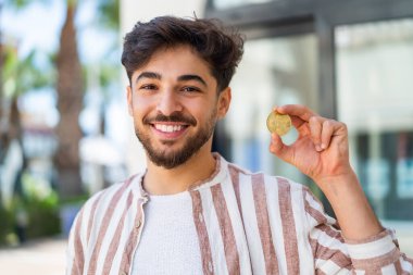 Yakışıklı bir Arap açık havada Bitcoin tutuyor, neşeli bir ifadeyle.