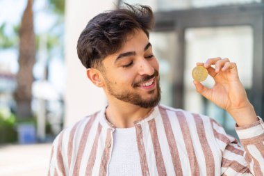 Yakışıklı bir Arap açık havada elinde mutlu bir ifadeyle Bitcoin tutuyor.